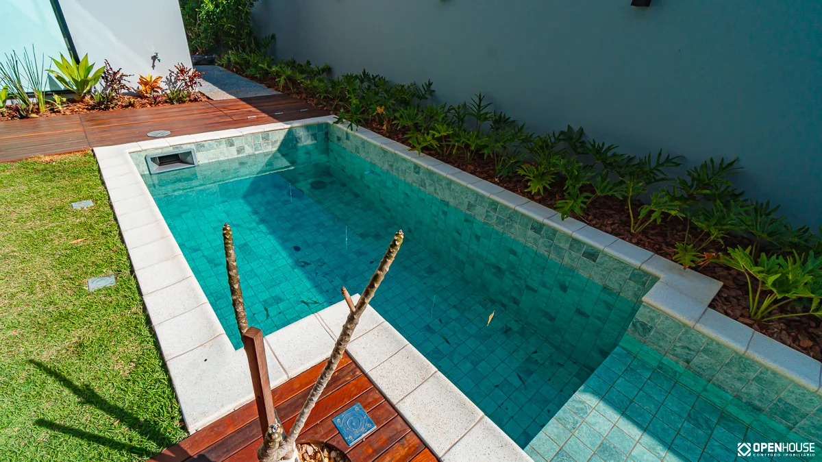 Piscina linear com área rasa contígua e deck em madeira junto aos panos de vidro da residência