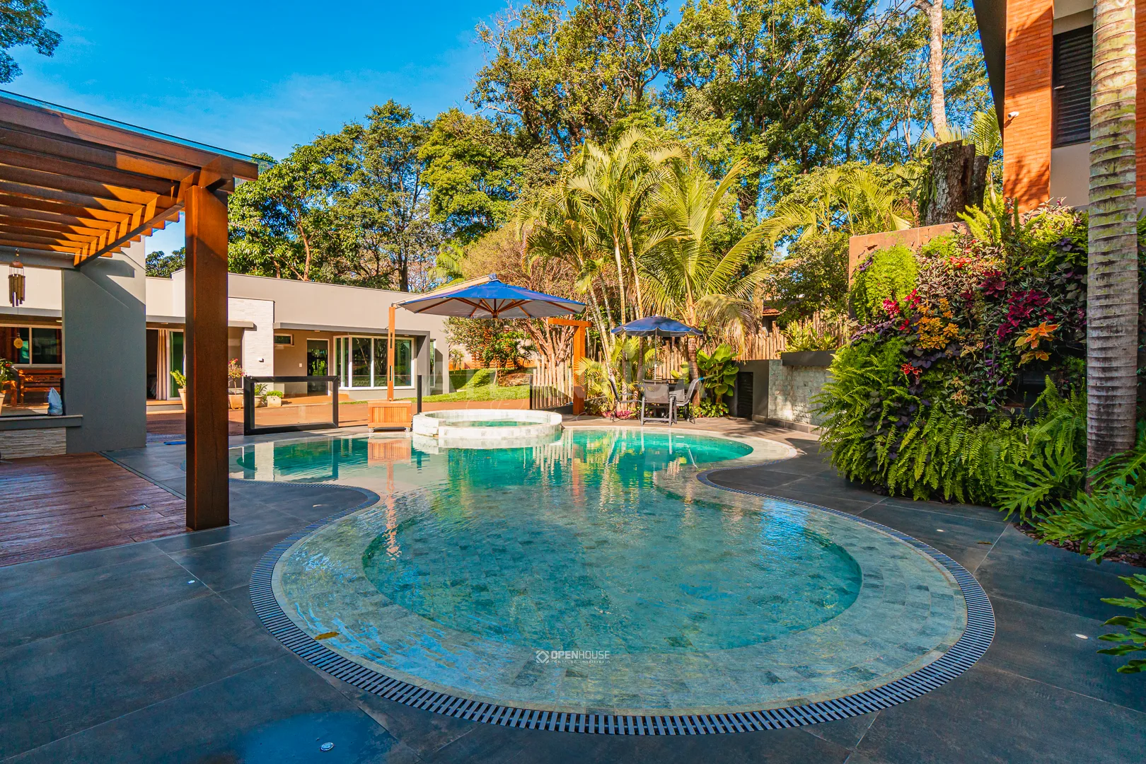 Visão geral do pátio com a piscina curva, spa circular, guarda-sóis e vegetação alta ao redor