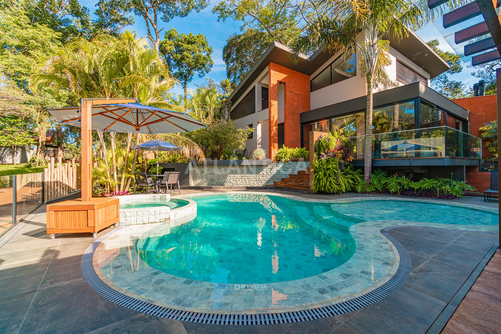 Piscina de contorno curvo junto ao guarda-sol e ao paisagismo, com a residência ao fundo