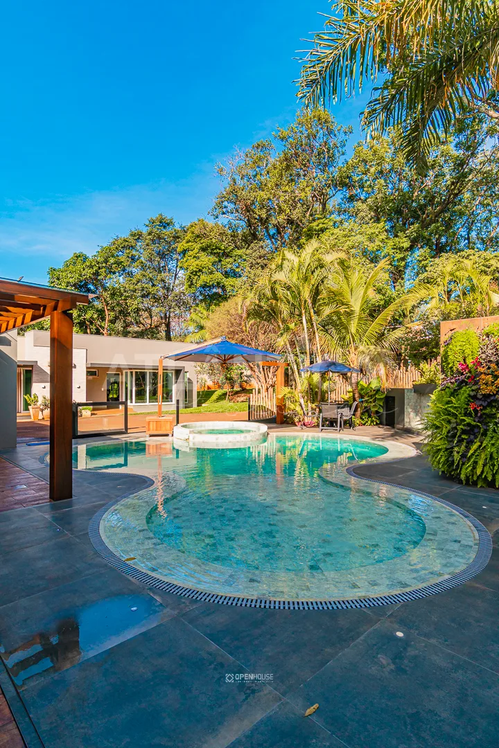 Visão ampla do jardim com árvores altas, guarda-sóis e a piscina de linhas orgânicas em primeiro plano