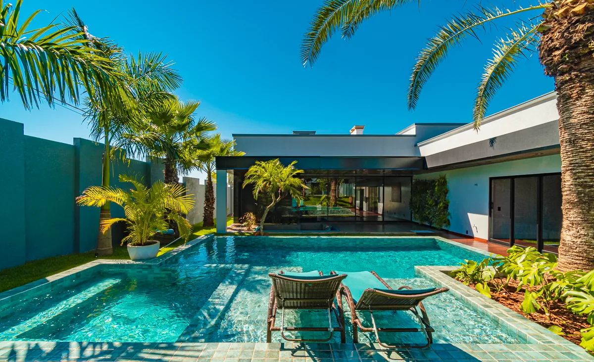 Piscina elevada revestida em pastilhas verdes com espreguiçadeiras sobre plataforma e casa térrea ao fundo