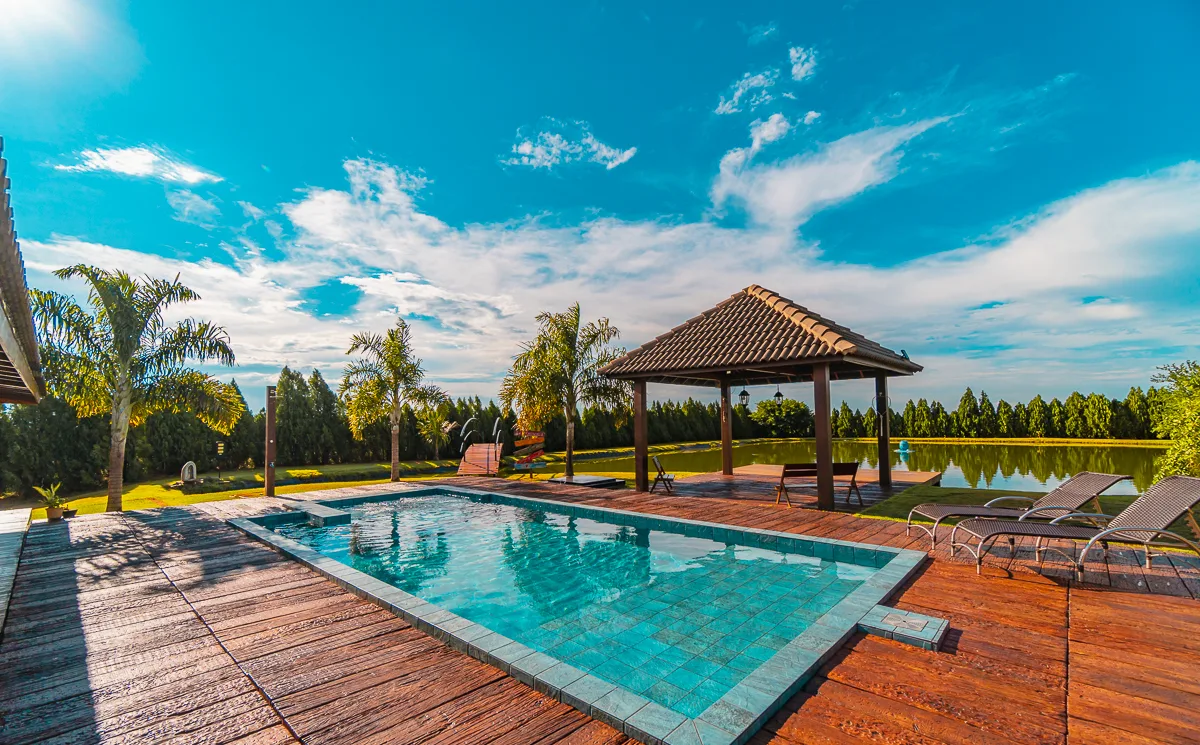 Piscina retangular com deck de madeira, gazebo em telha cerâmica e lago ao fundo, na sede da fazenda